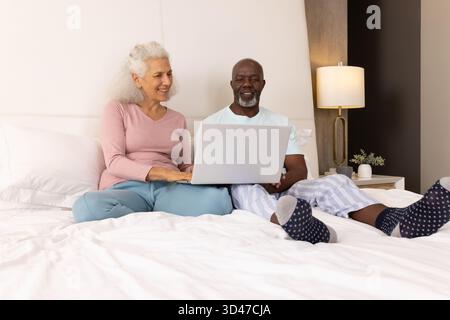 Typing senior couple sharing quiet morning on bed at home, with silver laptop and bedside lamp Stock Photo