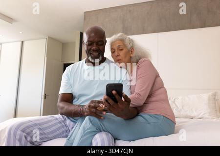 Smiling diverse senior couple looking at smartphone on bed in modern bedroom, with white linens Stock Photo