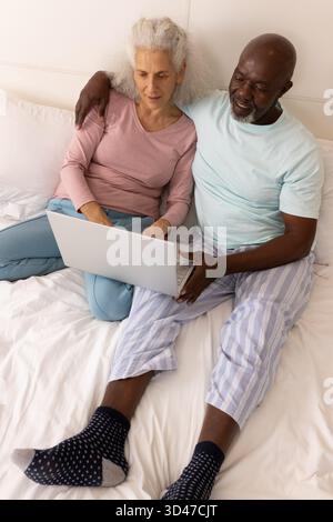 Senior couple using silver laptop while sitting on bed in bedroom at home, with white linens Stock Photo