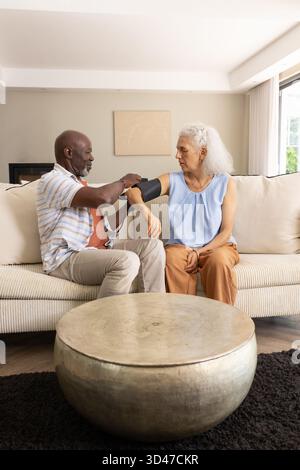Measuring blood pressure senior African American man assisting mature woman on sofa, with cuff Stock Photo