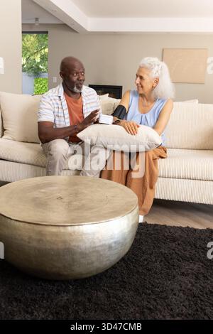 Using blood pressure cuff on Diverse senior couple sitting on sofa in living room, with pillow Stock Photo