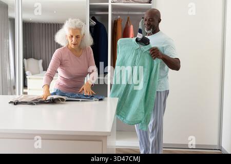 Organizing diverse senior couple folding jeans and holding shirt in walk-in closet, with countertop Stock Photo