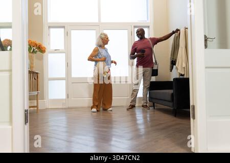 Chatting woman and senior African American man in home foyer, with mesh grocery bag and smartphone Stock Photo