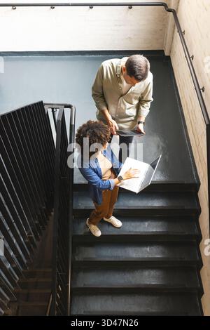 Collaborating Diverse coworkers reviewing digital files on office stairwell, with laptop and tablet Stock Photo