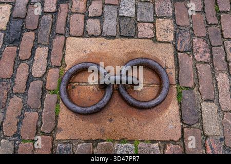 Two linked antique iron mooring rings set in granite, surrounded by wet, weathered cobblestones rustic maritime urban detail. Stock Photo
