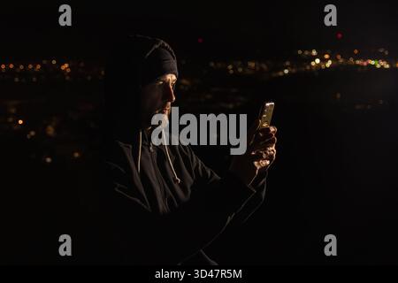 Man in hoodie sitting above Czech city and working on mobile phone at night. Hacker, remote working concept background Stock Photo
