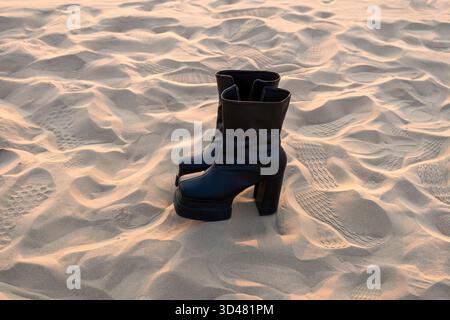 Doha, Qatar - October 30, 2025: A pair of stylish black high-heeled boots standing on soft desert sand during a serene sunset. Stock Photo