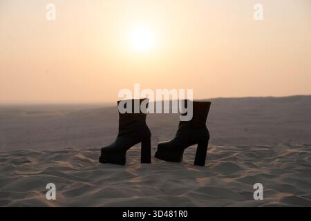 Doha, Qatar - October 30, 2025: A pair of stylish black high-heeled boots standing on soft desert sand during a serene sunset. Stock Photo