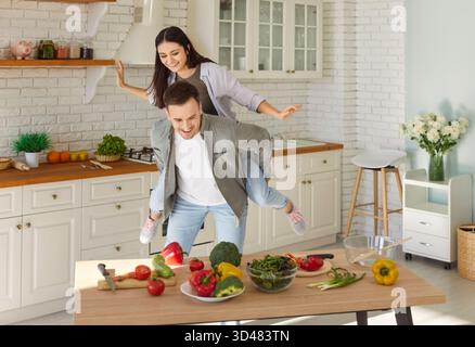 Funny playful piggyback existing couple happy fun in healthy amuse cooking salad in modern kitchen Stock Photo