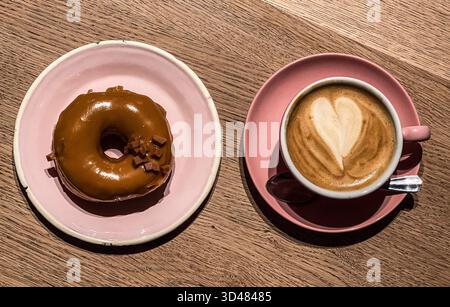 Chocolate covered ring donut on a pink plate alongside a cappuccino coffee, as served in Brammibal's Donuts, Berlin. The donuts are Vegan handmade don Stock Photo