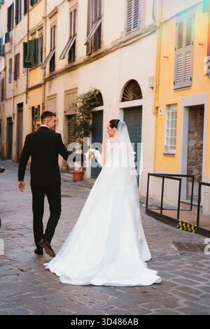Newly married couple walks hand in hand down a charming European street. The bride in an elegant dress and the groom in a suit share a joyful moment a Stock Photo