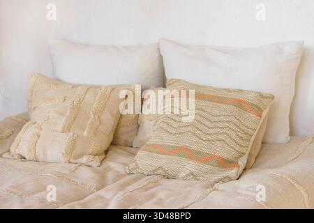 Decorative beige pillows, cushions with woven textures and natural linen covers arranged on cozy bed in bedroom interior. Warm tones creating peaceful Stock Photo
