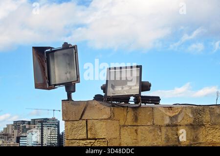 Two industrial floodlights mounted on a stone wall against a cloudy blue sky and modern city skyline on the horizon. Stock Photo