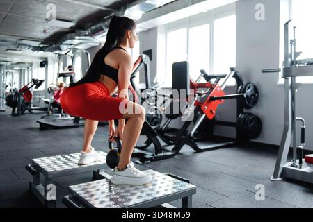 Active young female in red shorts and black top doing squat exercises with kettlebell at fitness center. Concept of self discipline and body care. Stock Photo