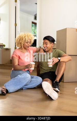Young diverse couple sitting on floor with coffee cups, enjoying new home together. Moving, relocation, new beginnings, relaxing, settling, residential || Model released Stock Photo
