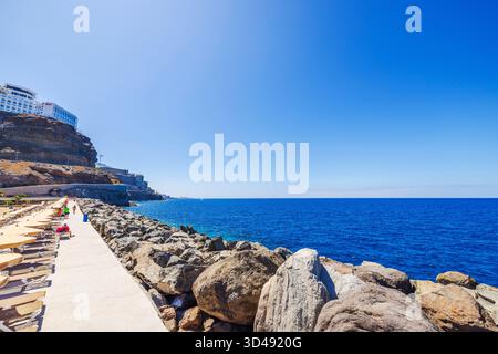 Sunny promenade near Amadores Beach with sun loungers and tourists walking along rocky coast on background of Atlantic Ocean and Riu hotel. Stock Photo