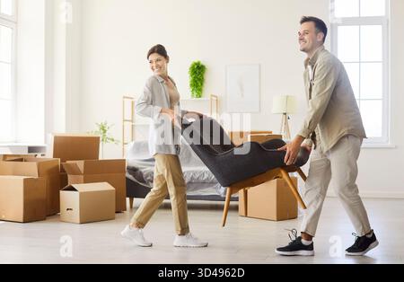 Smiling couple moving into apartment, carrying armchair among boxes and planning Stock Photo