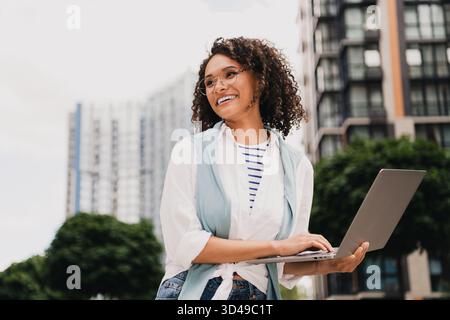 Young woman outdoors with laptop in urban city park wearing striped shirt and light vest smiling at work Stock Photo