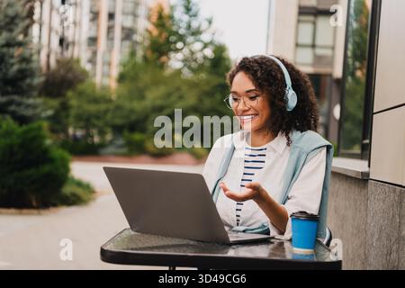 A smiling young woman with headphones works on a laptop at an outdoor cafe in an urban city setting Stock Photo