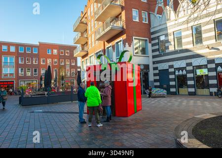Festive public plaza scene featuring a giant red gift box with green ribbon and small group of people nearby in center of Waddinxveen, Netherlands Stock Photo
