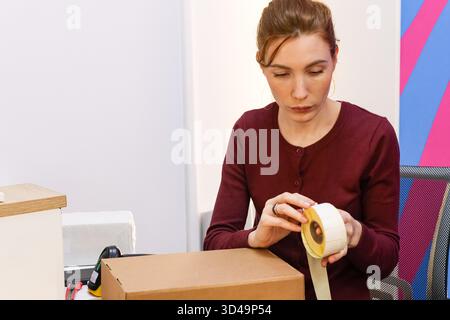 Woman sealing cardboard box with tape at delivery pickup point. Close-up of female worker applying adhesive tape to seal a brown parcel in a logistics Stock Photo