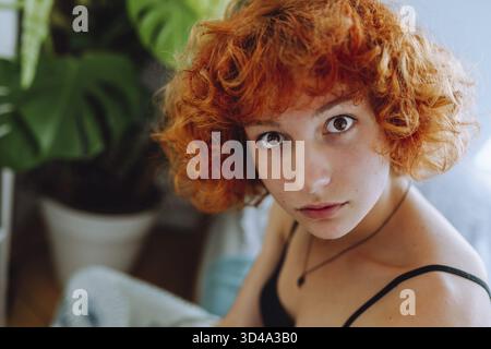 Portrait of young woman with curly red hair sitting on bed in bedroom using phone, monstera plant in background Stock Photo
