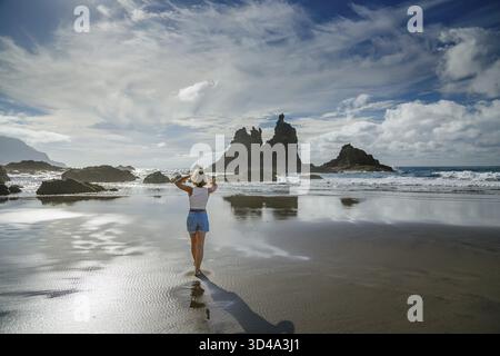 A woman standing barefoot on wet sand at Playa Benijo, Tenerife, with ocean waves gently touching the shore and dramatic rock formations in the backgr Stock Photo