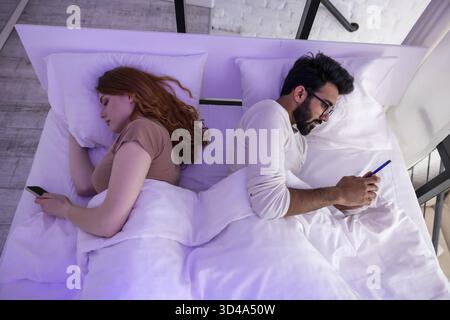 Young couple in bed using phone lying backs to each other Stock Photo