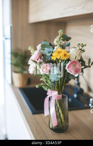 A beautiful bouquet of assorted flowers placed in a clear glass vase with a pink ribbon, set on a countertop against the backdrop of a modern kitchen Stock Photo
