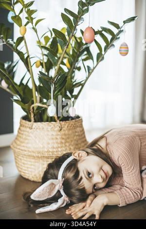 A smiling girl in a pink dress and fluffy bunny ears headband lying in front of decorated houseplant with festive Easter eggs Stock Photo