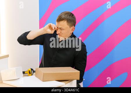 Man rubbing eyes at pickup counter with package. Tired worker at delivery pickup point rubs eyes near cardboard box on counter in colorful interior Stock Photo
