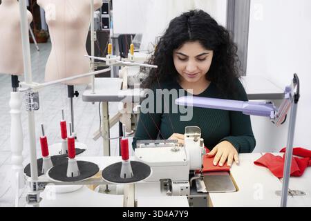 Young woman using professional overlock sewing machine in workshop. Equipment for edging, hemming or seaming clothes at tailors shop Stock Photo