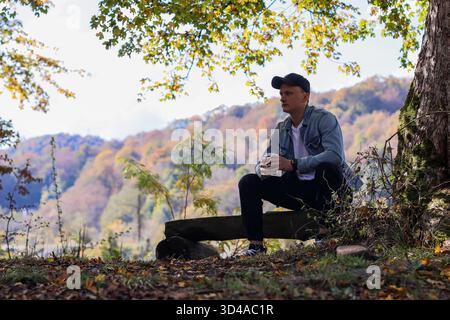 Man enjoying coffee in autumn mountain setting. Man wearing hoodie and jeans sits on log holding coffee cup surrounded by colorful fall leaves Stock Photo