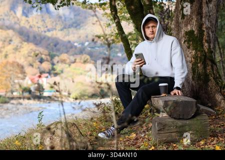 Relaxed man checking phone by river. Young man in white hoodie sits on log holding smartphone and coffee cup, overlooking autumn river and mountains. Stock Photo