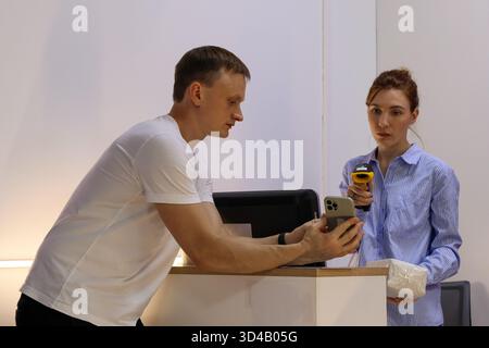 Worker scanning customer's smartphone at delivery pickup point. Woman in striped shirt holds scanner to man's phone over counter with monitor and pack Stock Photo
