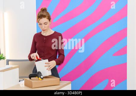 Woman using scissors on white package over brown box at counter. Worker in maroon top cuts into white parcel with red scissors beside scanner on woode Stock Photo