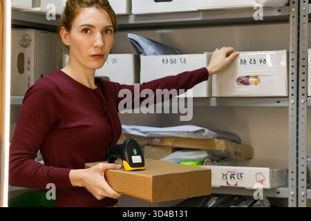 Woman holding box and scanner while reaching for storage bin. Female worker in maroon top holds cardboard box and barcode scanner Stock Photo