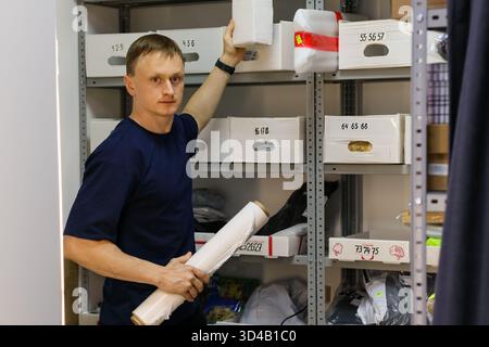 Employee holding roll and placing box on shelf. Young man holds packaging roll in one hand while placing white box on storage shelf with parcels. Stock Photo