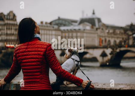 Paris, France 8 October 2024 Woman in red jacket with small Yorkshire terrier dog by the river. Travel lifestyle with pet, urban exploration, companio Stock Photo