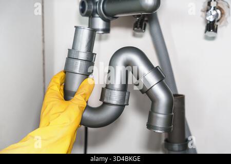 Person with rubber gloves detaches drainpipe beneath sink to fix clogged line and inspect for residue, Pipe cleaning, DIY plumbing Stock Photo