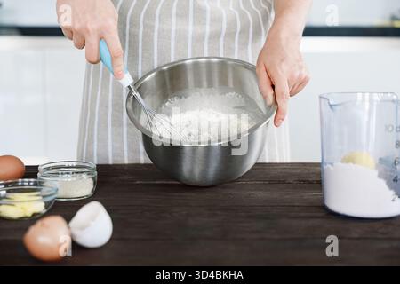 Baker in apron whisks flour with eggs, preparing dough for baking in metal bowl on wooden table, kitchen background Stock Photo