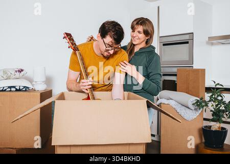 Man unboxes acoustic guitar while woman stands beside him smiling, surrounded by cardboard boxes and home goods, moving in new house Stock Photo