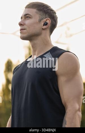 male athlete stands confidently on a sports ground with headphones on focused on his training as the sun sets highlighting his athletic build and determination. Stock Photo