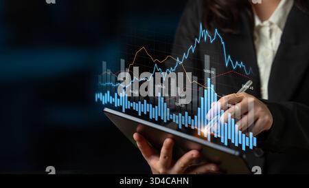A professional woman analyzes financial data using a tablet. Digital charts and graphs symbolize analytics and trends in a modern office setting Stock Photo