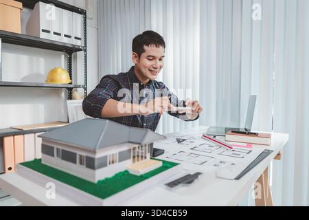 Young male architect working at office desk, drawing construction plans and using drafting tools, focused on building design while showing creativity Stock Photo