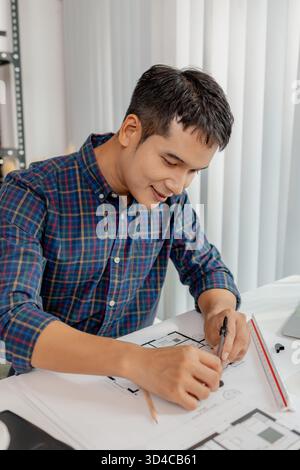 A young male architect is working with blueprints at his desk, using rulers and drawing tools to refine details. He appears focused and engaged, balan Stock Photo