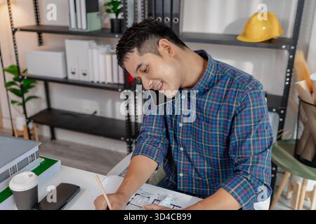 A young architect is multitasking at his desk, sketching blueprints, making phone calls, and reviewing notes. His workspace shows focus, productivity, Stock Photo