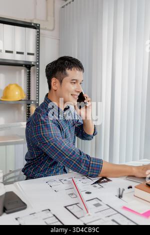 A young architect is multitasking at his desk, sketching blueprints, making phone calls, and reviewing notes. His workspace shows focus, productivity, Stock Photo