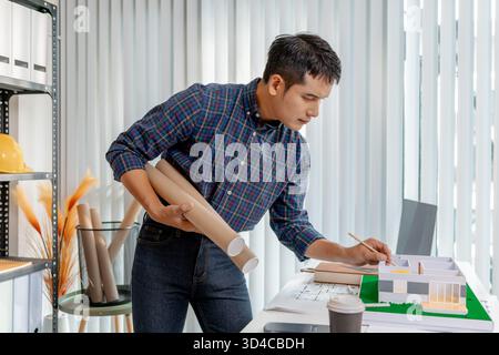 A young architect is working at his desk, reviewing blueprints, using a calculator, and checking a house model. He focuses on precision and detail whi Stock Photo