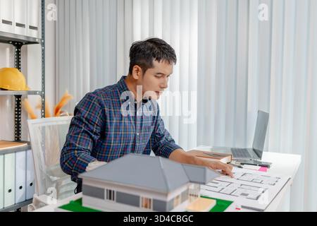 A young architect is working at his desk, reviewing blueprints, using a calculator, and checking a house model. He focuses on precision and detail whi Stock Photo